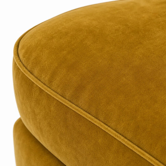 Close-up of a mustard yellow velvet or suede upholstered cushion with rounded edges and a visible seam, photographed against a white background.