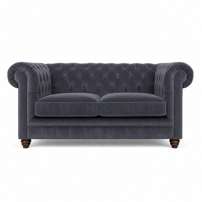 A classic Chesterfield loveseat sofa upholstered in dark slate blue velvet, featuring a tufted button backrest, rolled arms, two seat cushions, and dark wooden turned legs, shown against a plain white background.