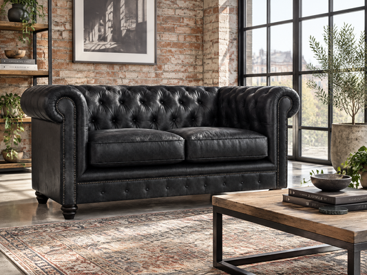 A black leather Chesterfield loveseat sofa with button tufting and nailhead trim, staged in a modern loft-style living room with a brick wall, large windows, a wooden coffee table, and a patterned rug.