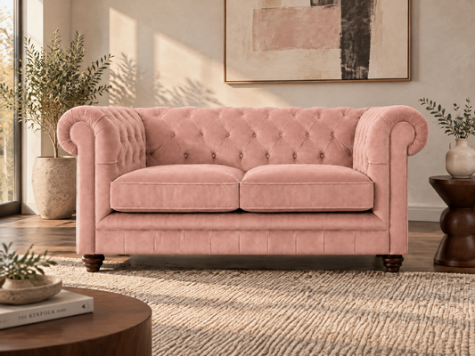 A pink tufted velvet loveseat sofa in a styled living room setting with a wooden floor, neutral rug, side table, and potted plants.
