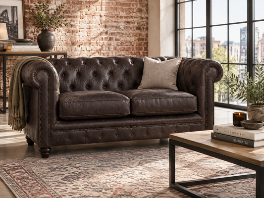 A dark brown leather Chesterfield loveseat sofa in a styled living room setting with a brick wall, large windows, a patterned rug, and a coffee table.