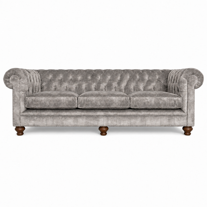 A classic Chesterfield-style three-seater sofa upholstered in textured gray velvet, featuring deep button tufting, rolled arms, and dark brown turned wooden legs, shown against a plain white background.