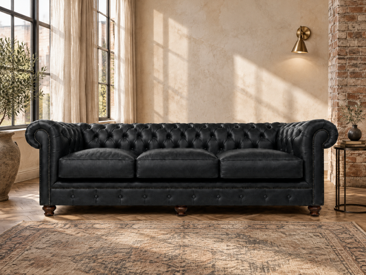 A black tufted leather Chesterfield sofa in a rustic, sunlit room with a brick column, textured wall, and patterned rug.
